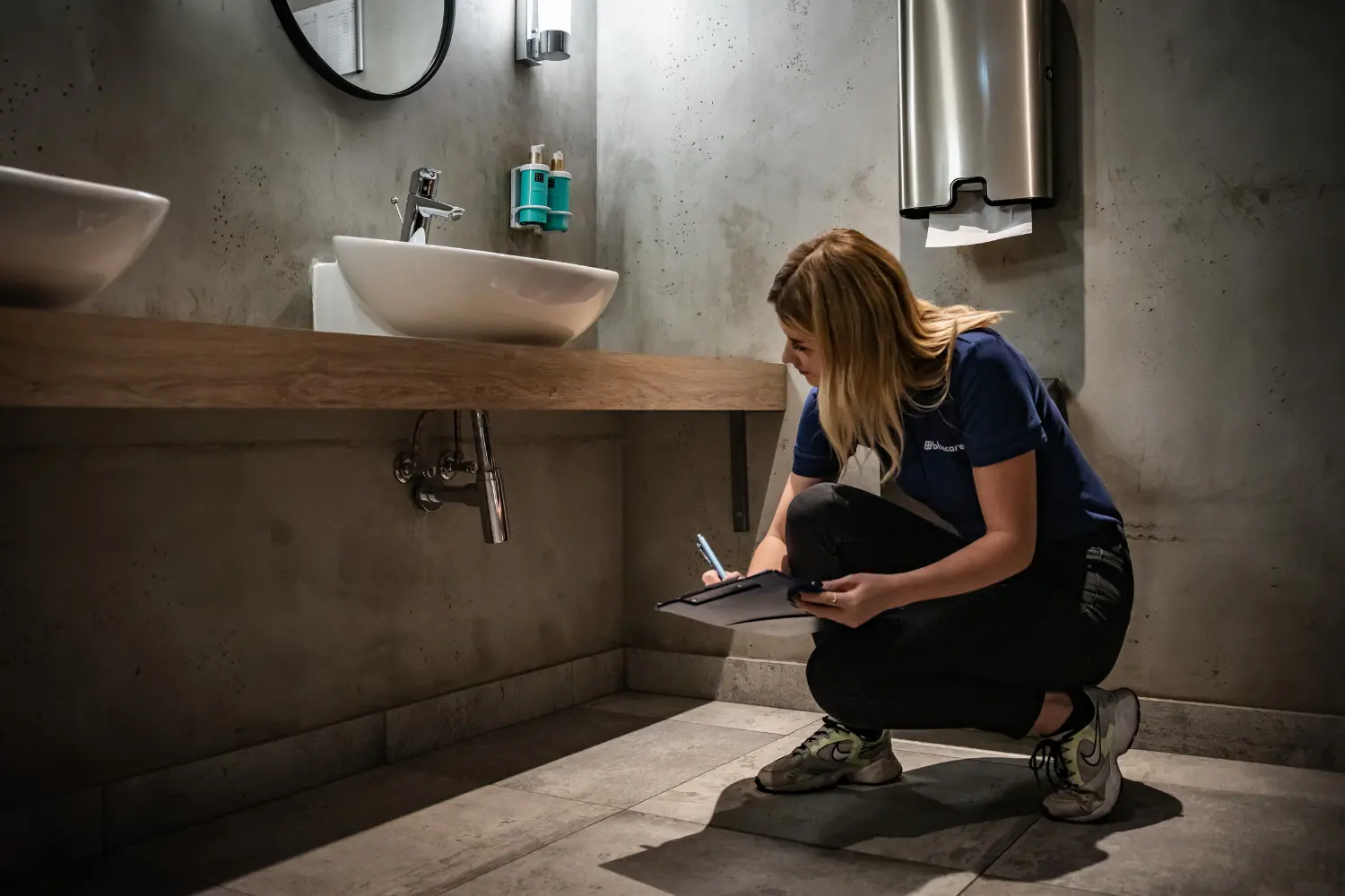 Technik audytu legionella kontrolujący i notujący stan instalacji wodnej pod umywalką w łazience.