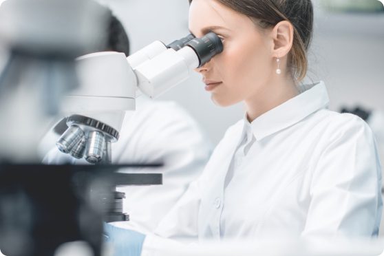 Młoda kobieta-laborantka w białym fartuchu prowadzi badania, patrząc przez mikroskop laboratoryjny- wykonuje badanie wody ze studni.