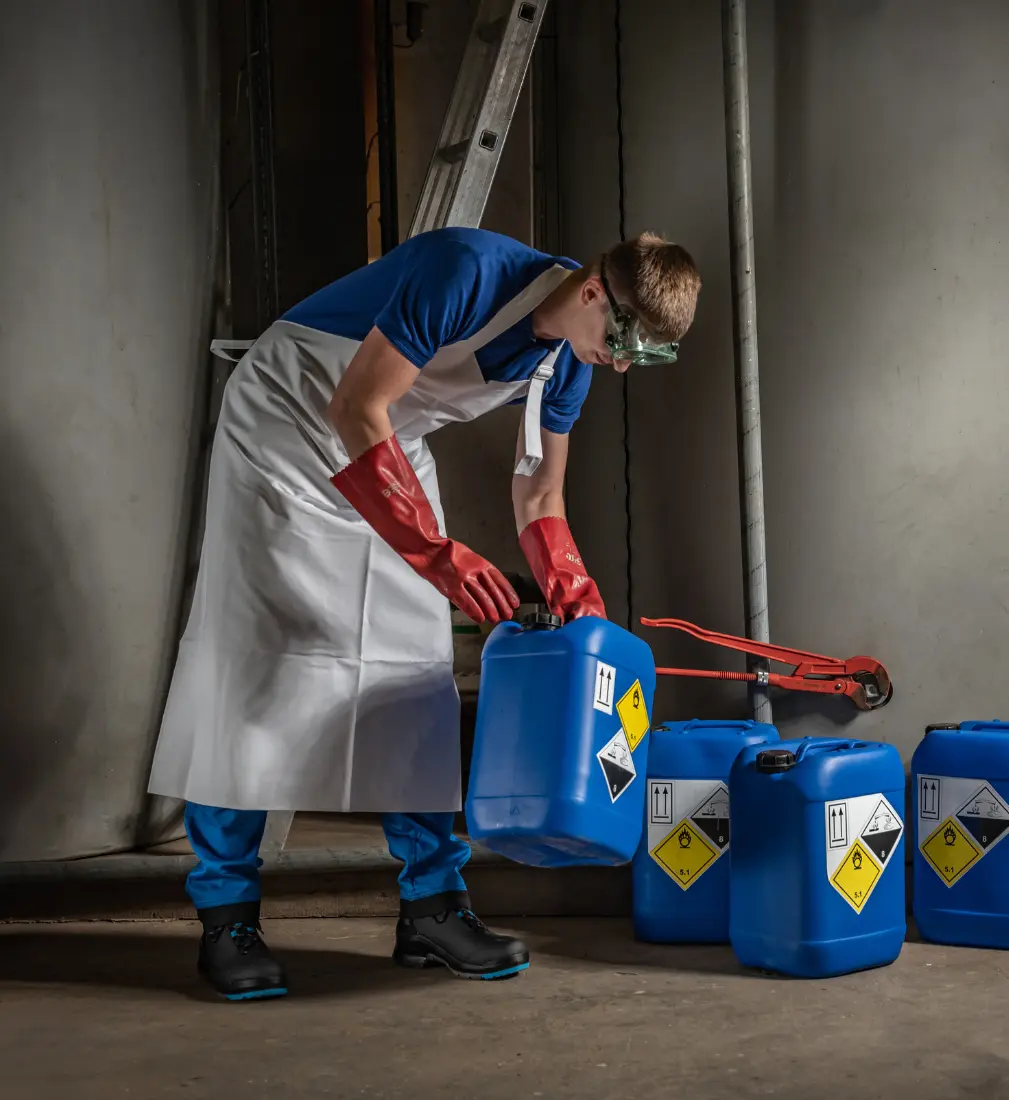 Pracownik w fartuchu i rękawicach ochronnych trzyma niebieski kanister z chemikaliami do dezynfekcji instalacji wodnych.