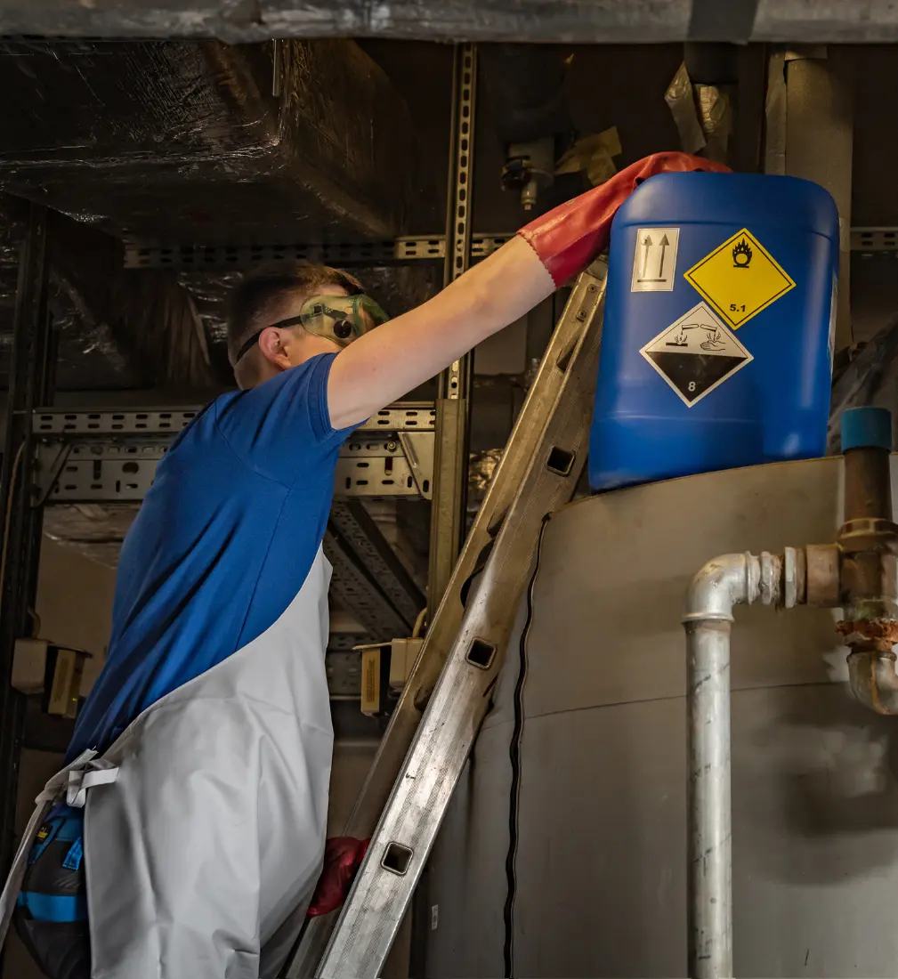 Technik w okularach i rękawicach BHP umieszcza niebieski pojemnik z chemikaliami na zbiorniku w trakcie czyszczenia instalacji podczas dezynfekcji chemicznej